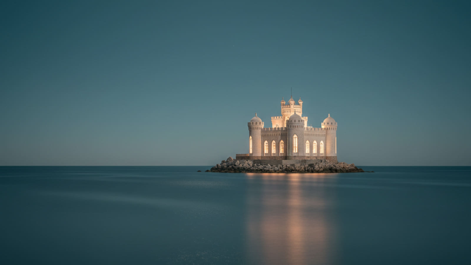 The castle at blue hour, warm light glowing from its windows above calm water.